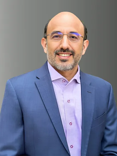 Un hombre con gafas, barba y calvo, vestido con una chaqueta azul y una camisa morada clara, sonriendo frente a un fondo gris degradado.